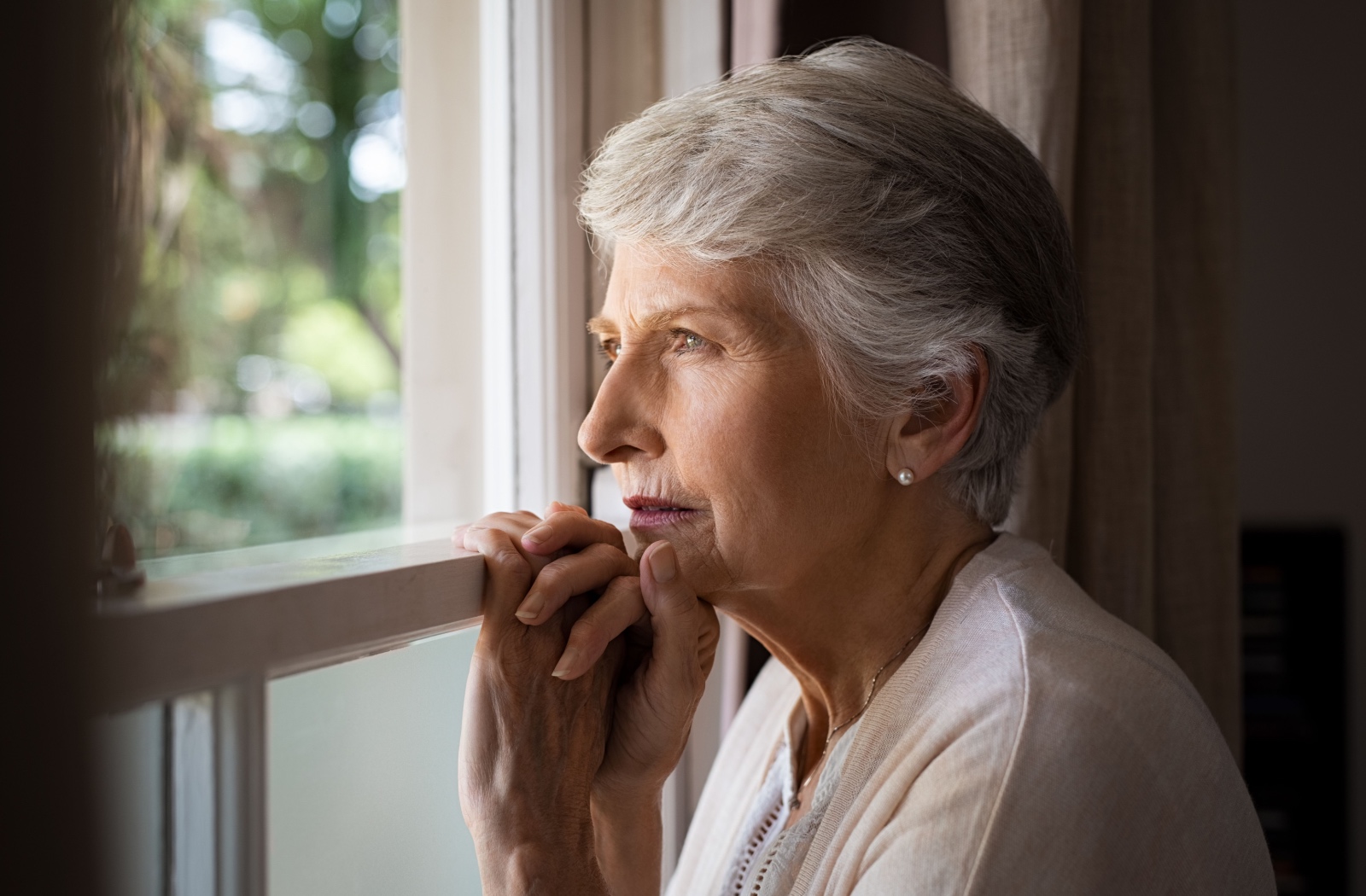 An older adult woman gazes solemnly out a window.