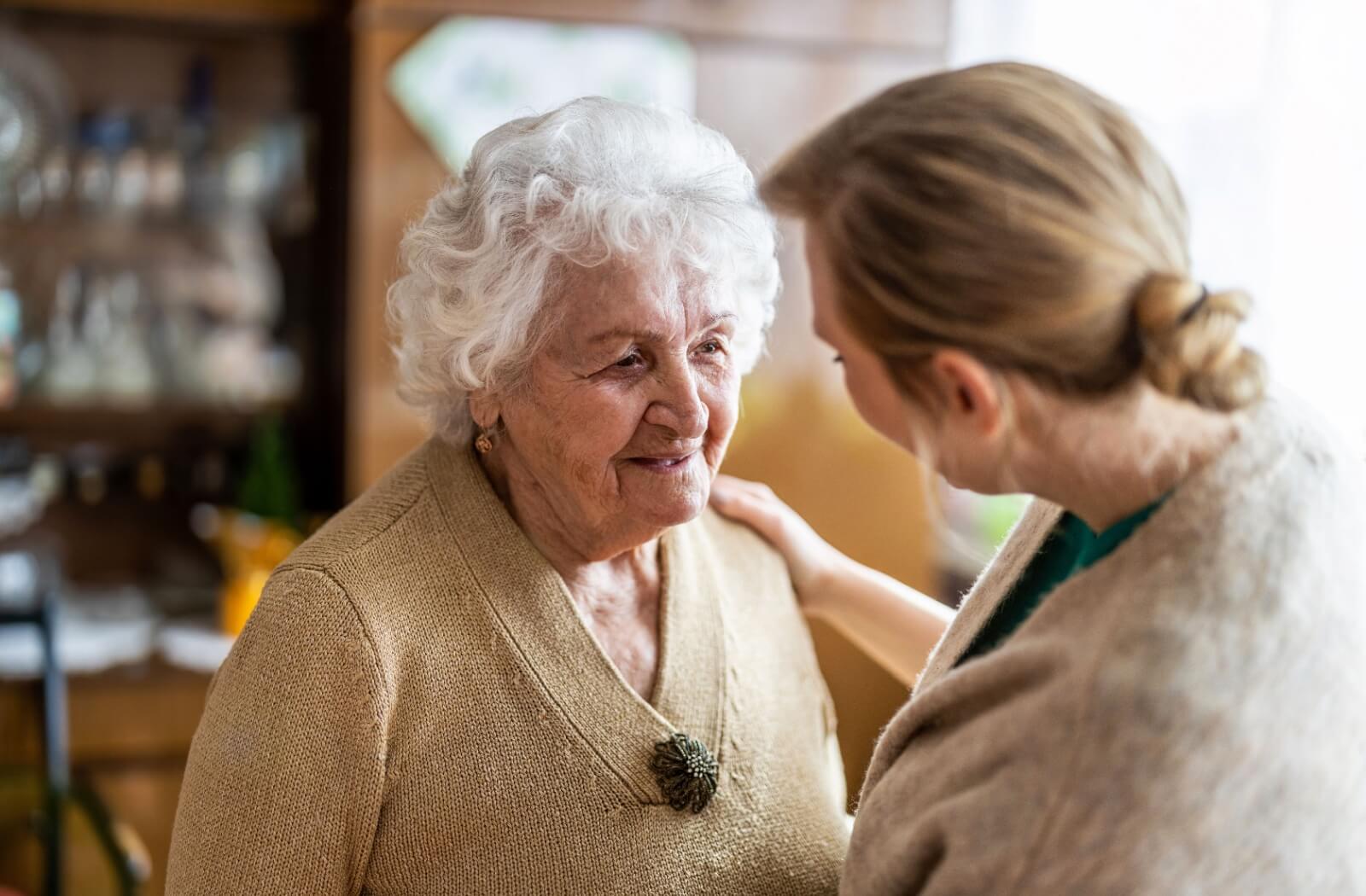An adult daughter places her hand on the shoulder of her mother as they converse.