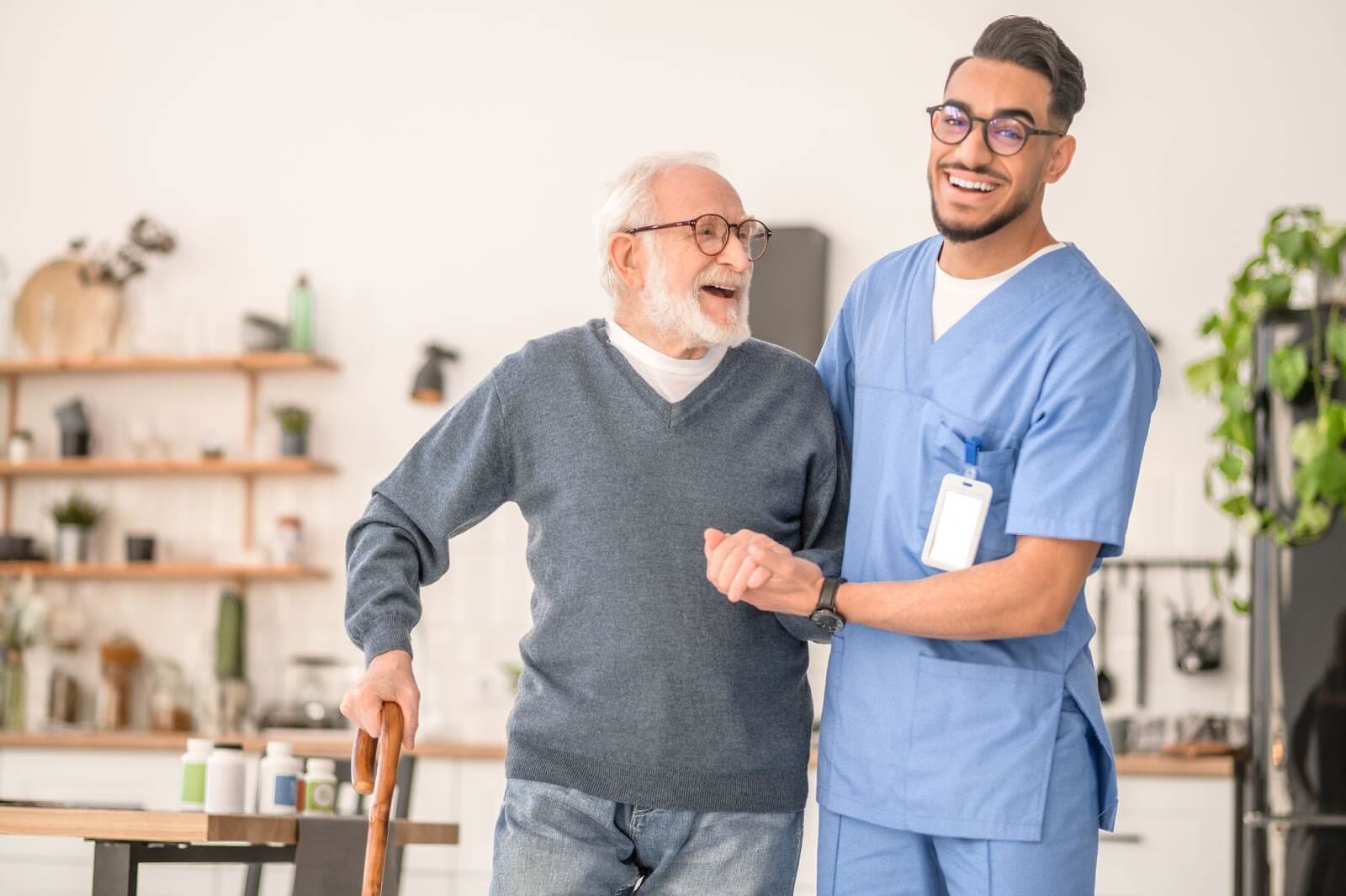 A smiling male caregiver in an assisted living community helps an older adult resident walk with a cane.