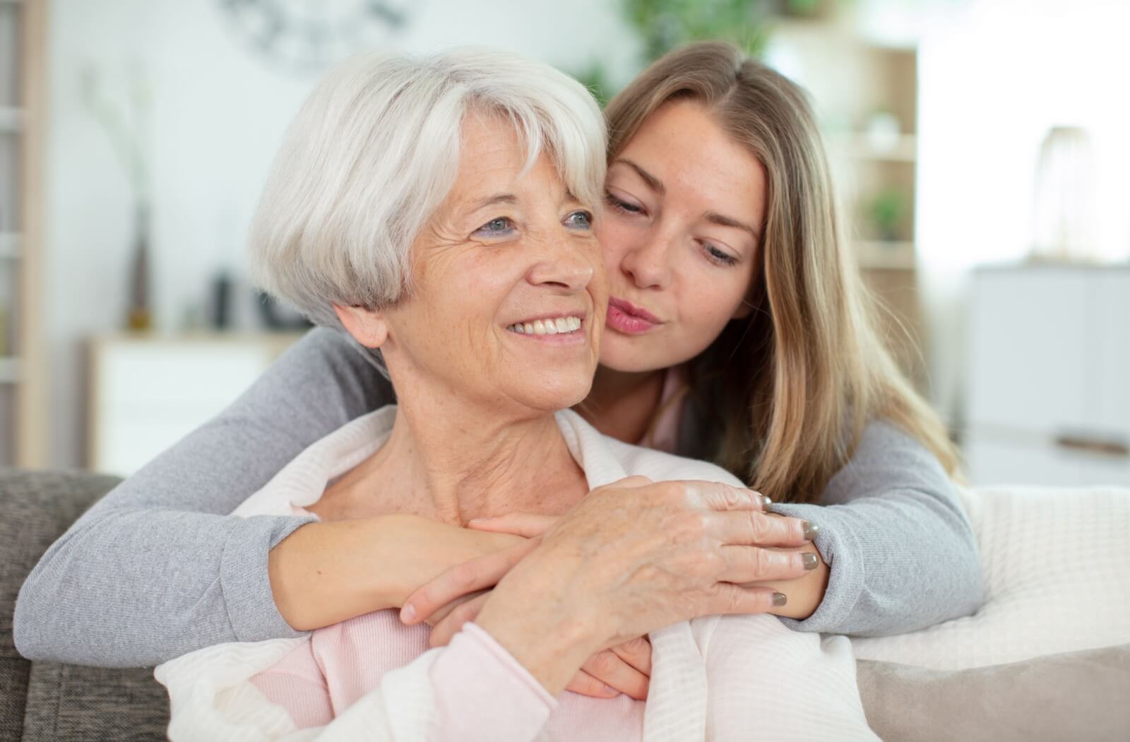 A daughter hugs her older adult mother from behind and kisses her cheek.