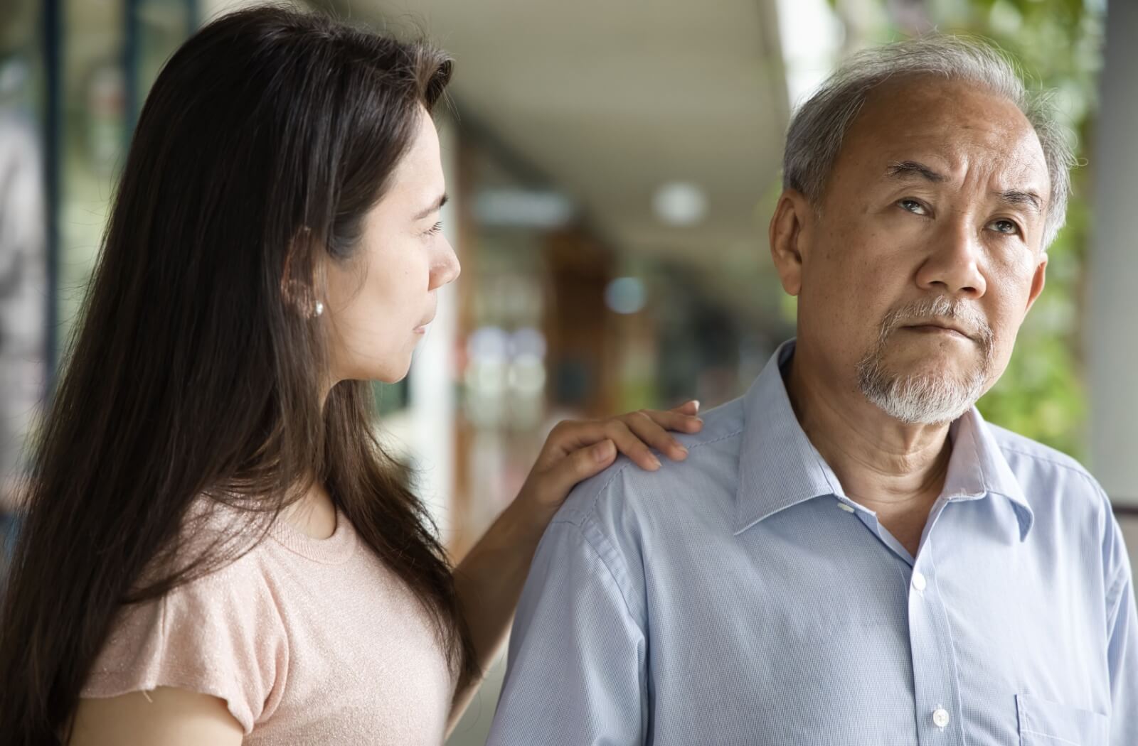 A person comforting an older adult outdoors by placing a hand on their shoulder while the older adult looks away with a distant expression.
