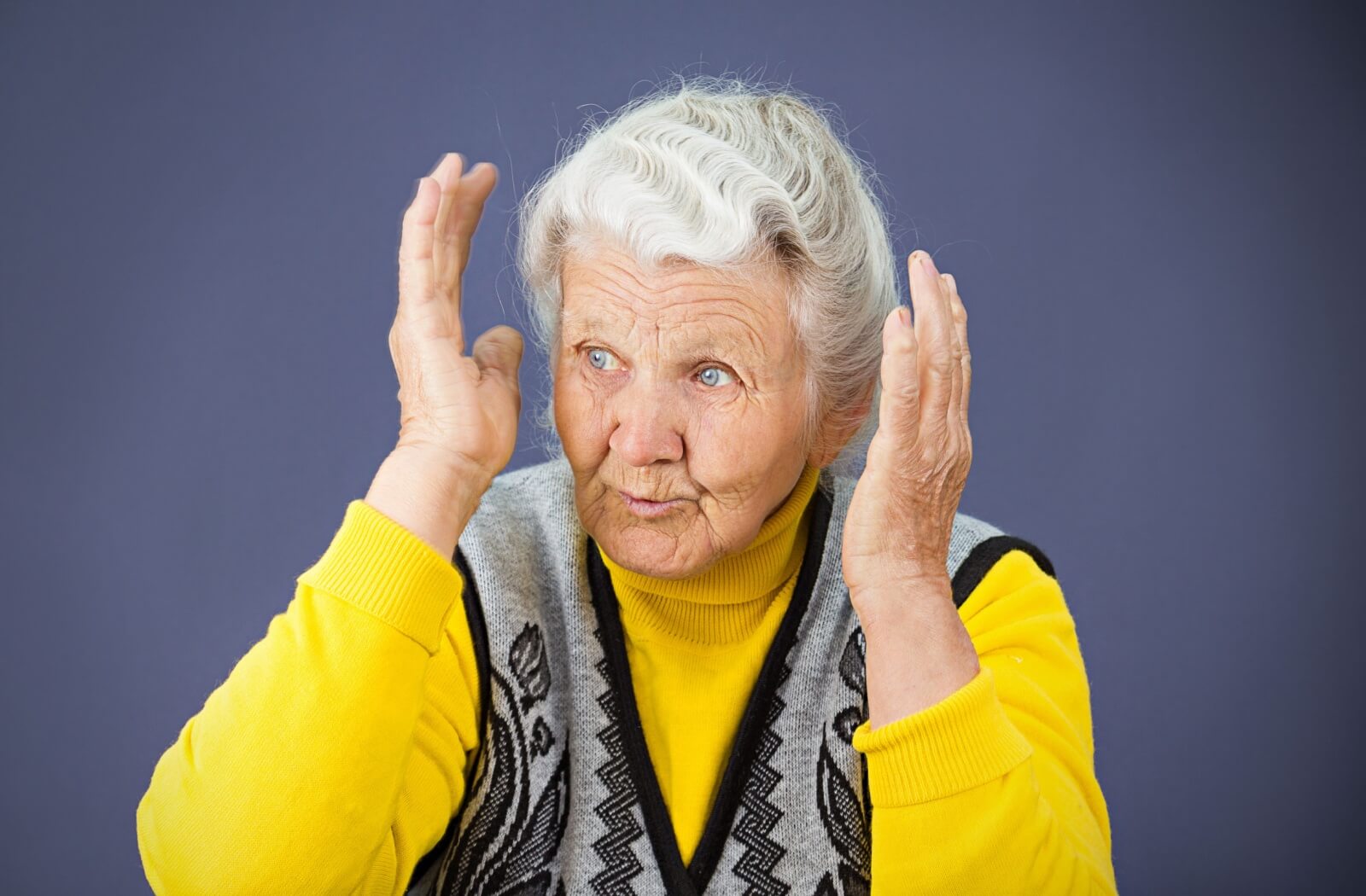 An older person with silver hair wearing a yellow sweater gestures expressively, appearing confused or distressed.