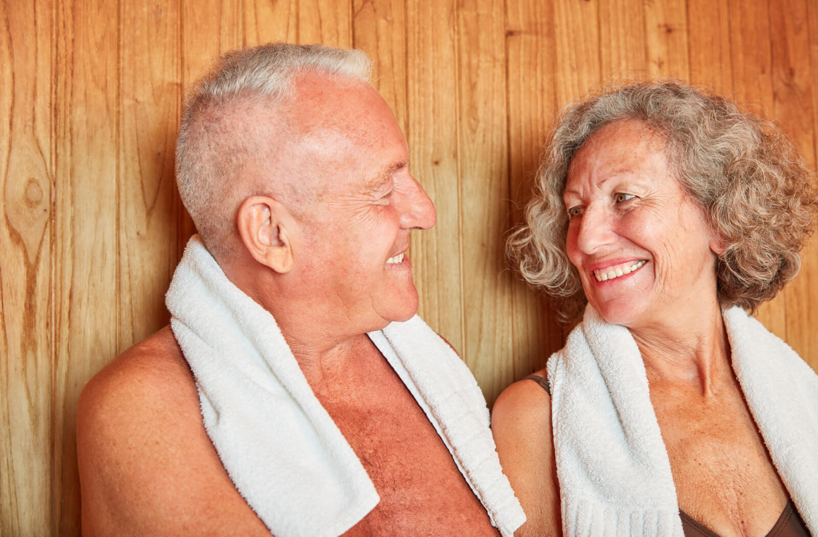 An older couple with towels around their neck smiling at one another while sitting in a sauna.