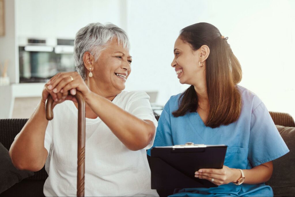 An older adult resident of a personal care home and a caregiver sit on a couch, look at each other, and smile.