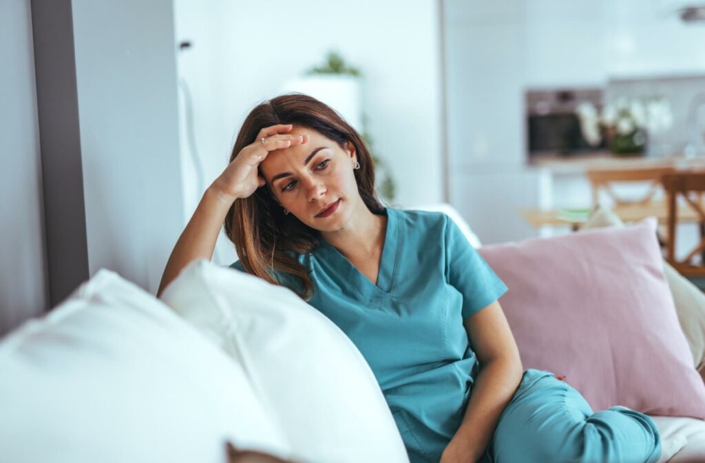 A young adult in blue scrubs sits on a couch and stares blankly at the floor, overwhelmed by caregiver burnout
